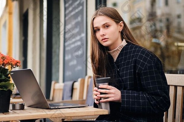 在室外咖啡馆休息时，迷人严肃的女孩自信地看着摄像机，手里拿着笔记本电脑