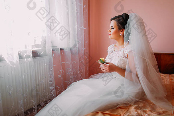 美丽的新娘坐在靠近窗户的床上，梦想着婚姻生活，手里拿着可爱的小结婚花束