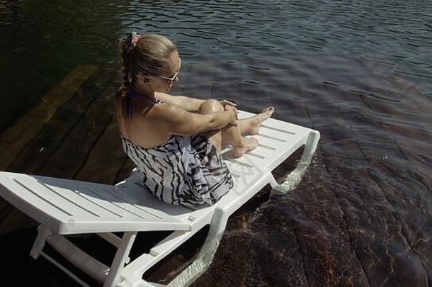 那个女人躺在日光浴床上，戴着太阳镜，披着波西米亚丝绸披肩。女孩在洪水淹没的木头水下码头上休息。湖里的路面被水覆盖着。