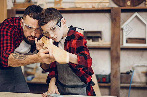 父亲身份，爱好，木工，木工和人的概念-父亲和小儿子戴着防护眼镜测试手工制作的木剑光滑冲浪