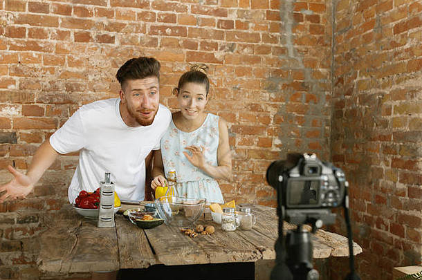 Vlogger和blogger自由职业，食品概念。一对年轻的高加索人夫妇在厨房的砖墙上用专业摄像机一起做饭，并为vlog和社交媒体录制现场视频。