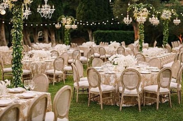 婚礼餐桌招待会。在绿色草坪上，为客人提供优雅的餐桌，带有图案的奶油桌布，挂着花环和枝形吊灯