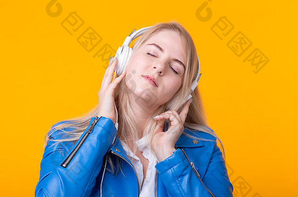 可爱、积极的年轻金发女孩的肖像，戴着耳机，穿着蓝色皮夹克，在黄色背景上摆姿势，听着最喜欢的音乐。概念