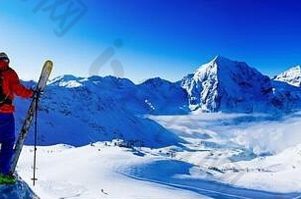 登山者背着滑雪板在雪山上休息。背景是蓝蓝的天空和灿烂的太阳，以及伊塔州南蒂罗尔的奥特勒