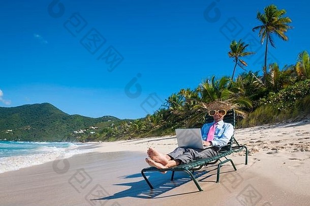 这位戴着草帽、戴着巨大太阳镜的商人在一张沙滩椅上用笔记本电脑打字，这张椅子在一个空荡荡的热带海滩的岸边远程工作