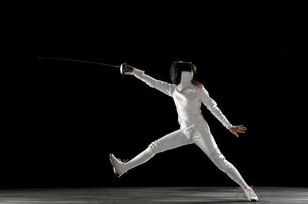 目标。身穿击剑服装的少女，手拿剑，黑色背景下被隔离。年轻女模特在运动、动作中练习和训练。空间。运动、年轻、健康的生活方式。