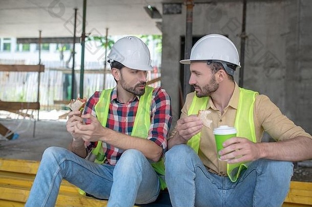 穿着黄色背心和头盔的建筑工人坐在木板上，一边吃三明治一边喝咖啡，一边聊天