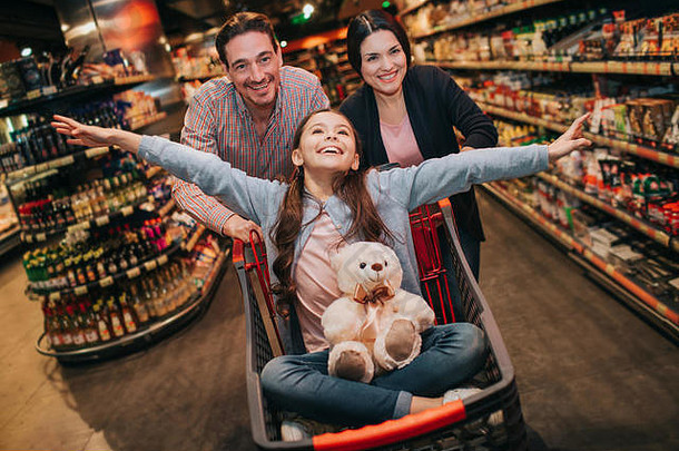 年轻的父母和女儿在杂货店。那个顽皮的女孩把玩具熊放在膝盖上。她假装在飞。父母站在后面，用手推手推车