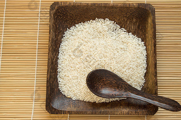 深色木盘上的生白米，带勺子，竹垫背景，俯视图，素食主义，有机食品，世界粮食日