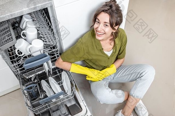 一位快乐的家庭主妇坐在洗碗机旁，家里厨房里放着干净的盘子。简单的家庭工作与厨房电器的概念
