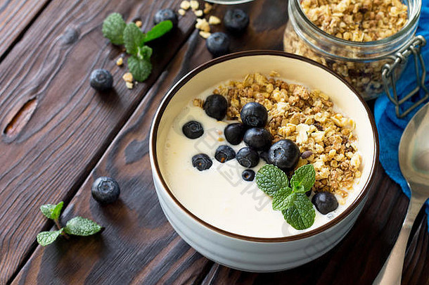 健康早餐放在碗里，烤格兰诺拉麦片、新鲜蓝莓和酸奶特写镜头放在厨房木制的乡村餐桌上。