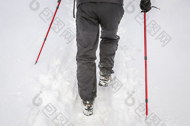 徒步旅行者双腿的后视图，带着登山杖行走在积雪深的小路上。
