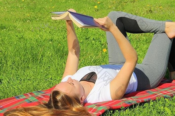一个穿着白色T恤、留着长发的年轻漂亮的白人女孩躺在一块<strong>红色格子</strong>布上，躺在绿草上，躺在草坪上看书。
