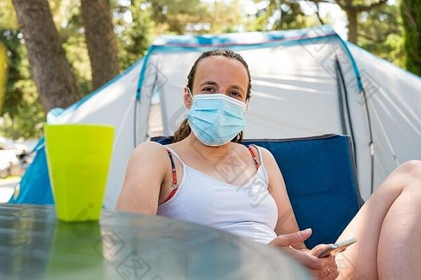 在一个度假胜地，一名戴着医用面罩的妇女坐在露营帐篷前。
