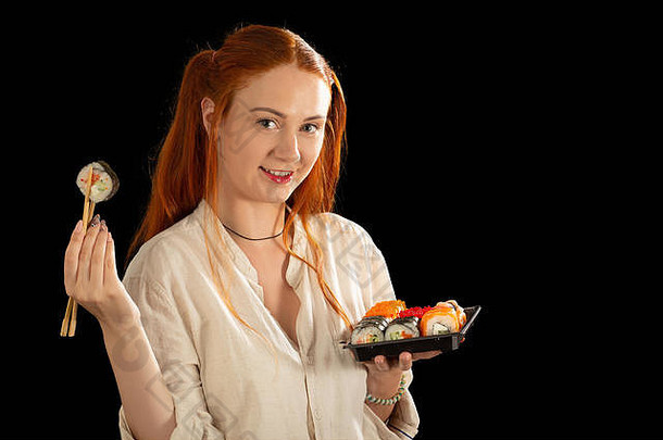 快乐的红发年轻女子在黑色背景下看着相机展示寿司