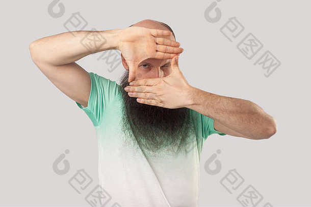 <strong>专注</strong>的中年秃头男子的肖像，留着长胡子，穿着浅绿色t恤，站在那里，做着作物构图的姿势，用手看着。在里面