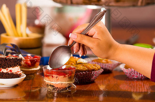 美味的慕斯，上面覆盖着草莓酱，放在小玻璃杯里，用手抓着勺子咬，蛋糕片和松饼背景，糕点概念