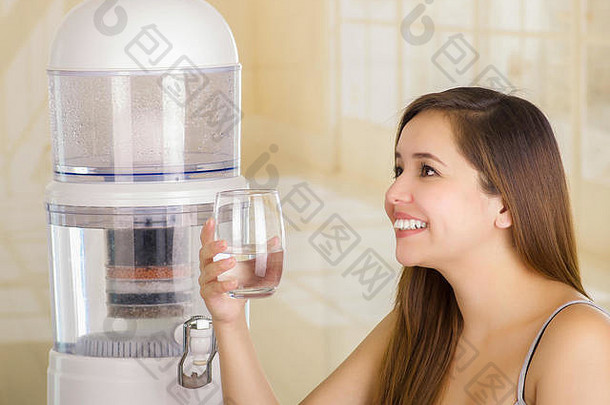 一个美丽的微笑着的女人拿着一杯水，厨房<strong>背景</strong>上有一个<strong>净水器</strong>的过滤系统