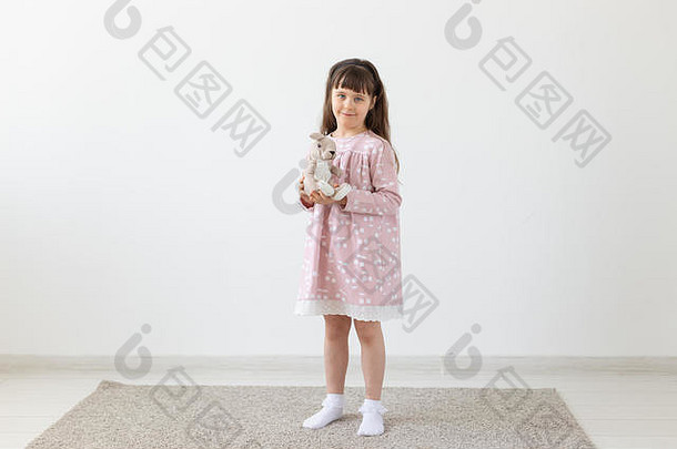 在白色的房间里，一个穿着粉红色连衣裙，带着玩具的小女孩
