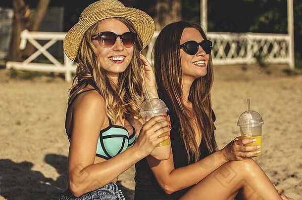 夏日快乐时刻。两个年轻漂亮的女人穿着泳衣，一边喝着柠檬水，一边微笑着在海滩上玩。