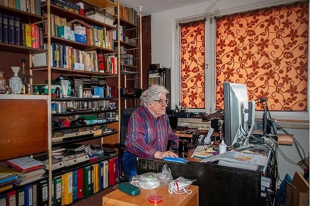 白发老人在家里用台式电脑工作，一片狼藉。书架上摆着许多书和抗病毒口罩