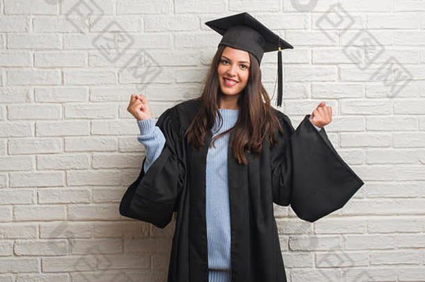 年轻的黑发女子站在白砖墙前，身穿毕业生制服，高声尖叫，自豪地庆祝胜利和成功，非常激动，加油