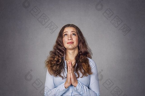 一名年轻女子在工作室灰色背景上独立祈祷的特写肖像。穿着蓝色正式衬衫，留着长波浪卷发的模特女孩。特写端口