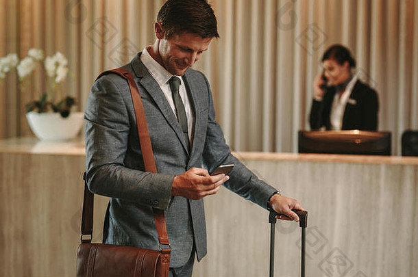 快乐的商人在酒店大厅使用手机。男客人带着电话和手提箱来到酒店。