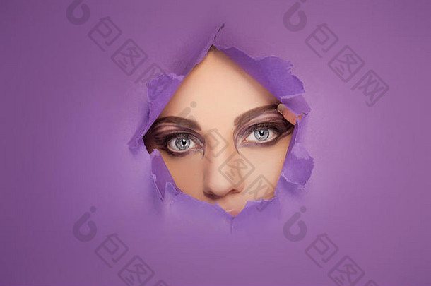 美丽的眼睛和富有创意的眼妆，透过紫色纸上的一个洞看的女人
