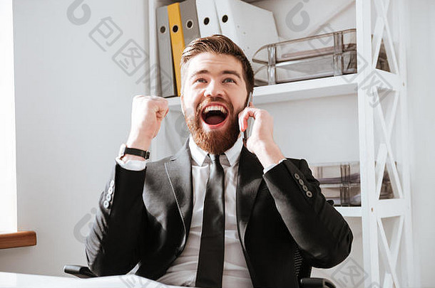 快乐的商人坐在办公室里，一边打电话一边做赢家的手势。