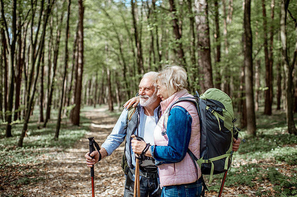 一对可爱的老年夫妇背着背包和<strong>登山杖</strong>在森林里拥抱。退休后积极生活方式的概念
