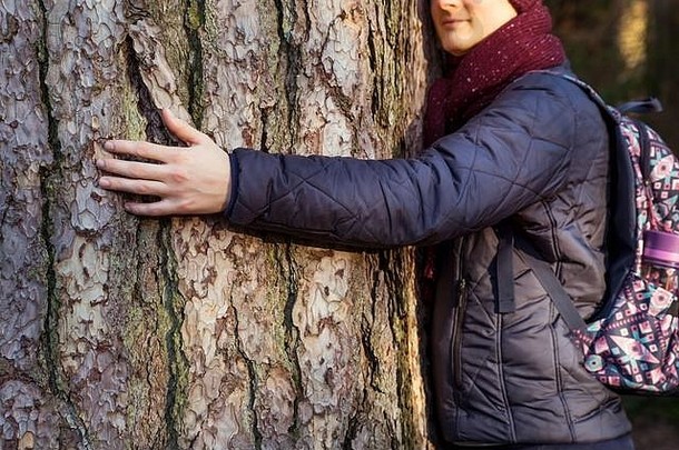 在森林里，一位年轻的自然爱好者背着背包，抱着一棵大树，脸上带着平静和微笑。专注于面部轮廓模糊的手部