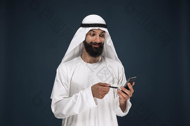 深蓝色工作室背景上的阿拉伯沙特商人肖像。男子使用智能<strong>手机支付</strong>账单、网上购物或。商业概念、金融、面部表情、人类情感。