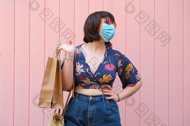 年轻的高加索人女人穿脸面具太阳镜持有购物袋粉红色的背景冠状病毒购物概念
