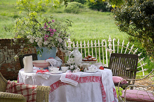郊野花园的草坪，桌上放着盛夏的鲜花，还有水壶和碗，还有镶着红边的白色亚麻布