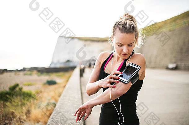 年轻的运动型女跑步者站在外面的海滩上，使用智能手机。