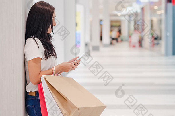 黑色星期五，一名妇女站在购物中心的背景上，一边使用智能手机，一边拿着购物袋。水平图像。