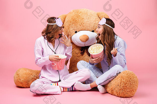 快乐漂亮的女孩穿着睡衣坐着大泰迪熊吃爆米花孤立的粉红色的背景