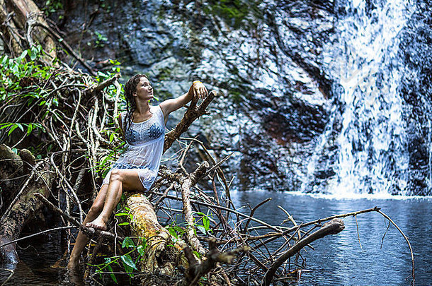年轻女子坐在森林瀑布池塘附近的树枝上。