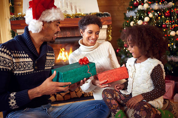 微笑的非裔美国人父母给他们的小女儿送圣诞礼物，每个人都坐在圣诞树前的地板上