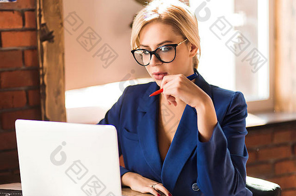 一位年轻漂亮的金发女商人的特写照片，戴着眼镜，穿着时髦的蓝色夹克，在咖啡馆里用白色笔记本电脑工作，手里拿着笔，<strong>严肃认真</strong>