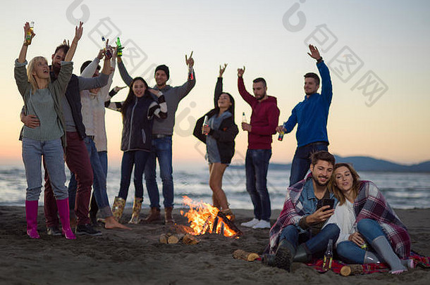 这对夫妇在秋季海滩派对上使用手机，和朋友一起喝啤酒，玩得很开心