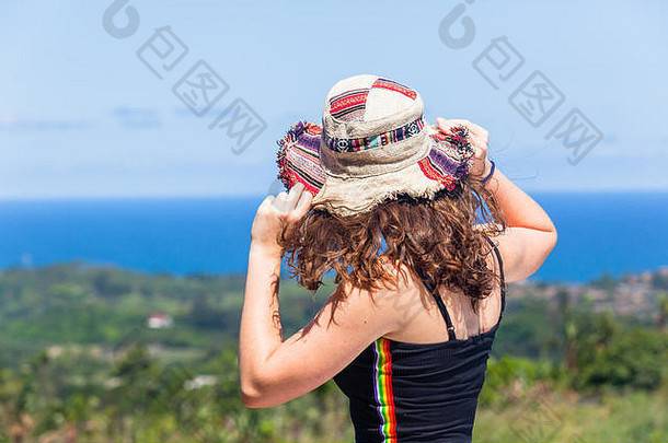 年轻少女度假特写镜头背肩后手拿帽子从风中眺望遥远的蓝色海洋和绿色的夏日