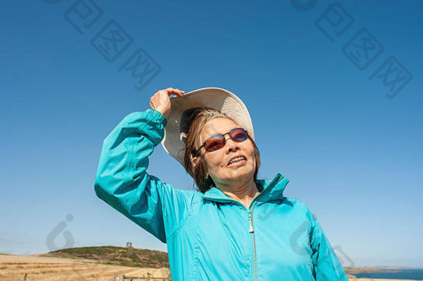 有魅力的成熟亚洲女士穿着蓝色的休闲服，帆布帽，太阳镜。用面部表情仰望蓝天进行<strong>解读</strong>。