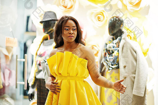 穿着黄色睡衣的时尚非洲裔美国妇女在商场里与模特儿对着陈列柜摆姿势。