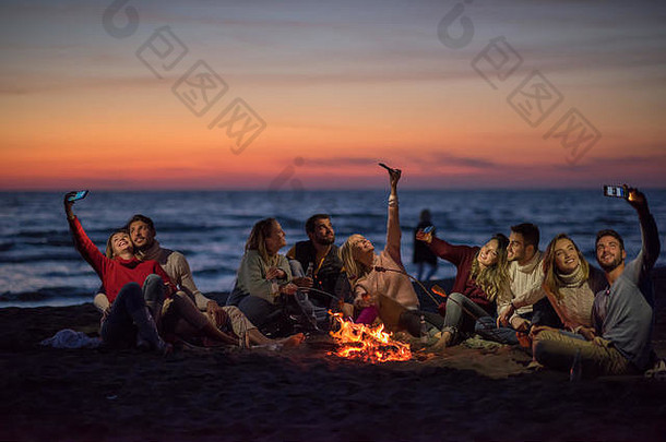 在秋天的海滩派对上，一群朋友在篝火旁用手机喝啤酒，玩得很开心