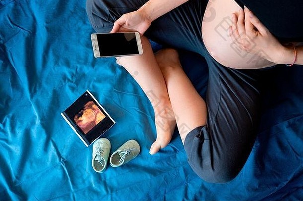 一个穿着婴儿鞋坐在蓝色床上看电话的怀孕女孩