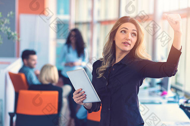 在办公室里，快乐的女商人手持数字平板电脑站在同事面前，在后台讨论