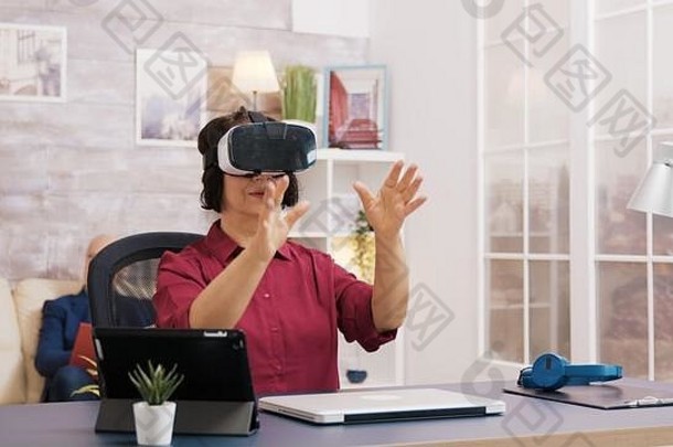老年妇女在客厅使用虚拟现实护目镜。坐在背景沙发上的老人。