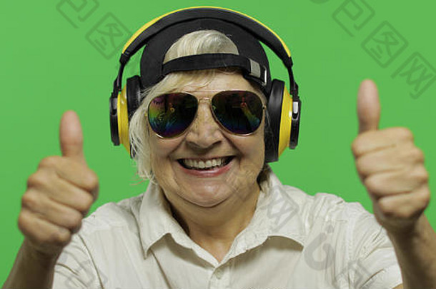 一位上了年纪的妇女戴着耳机听音乐。竖起大拇指。戴着墨镜和帽子的快乐老奶奶。放置您的徽标或文字。色度键。绿色屏幕背景